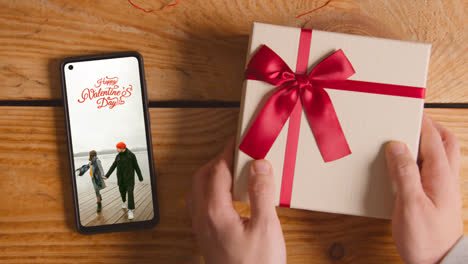 Close-Up-Of-Hand-Putting-Valentine's-Day-Gift-Next-To-Mobile-Phone-With-Valentines-Message-On-Screen